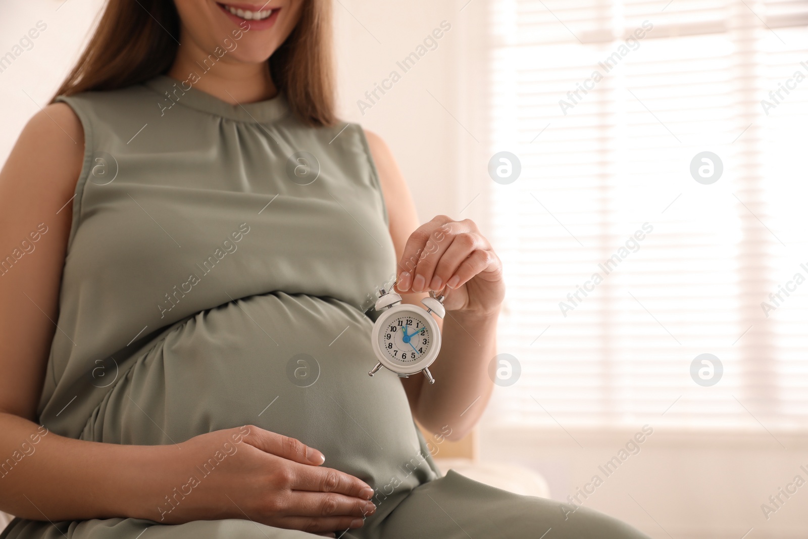 Photo of Young pregnant woman holding alarm clock near her belly at home, closeup. Time to give birth