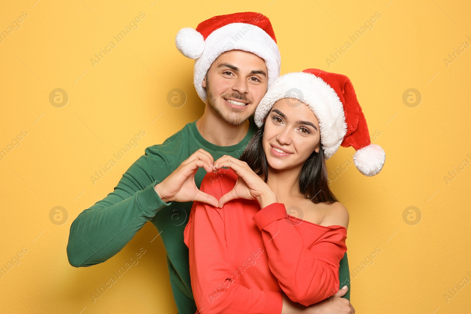 Photo of Young happy couple with Santa hats putting hands together in shape of heart on yellow background. Christmas celebration