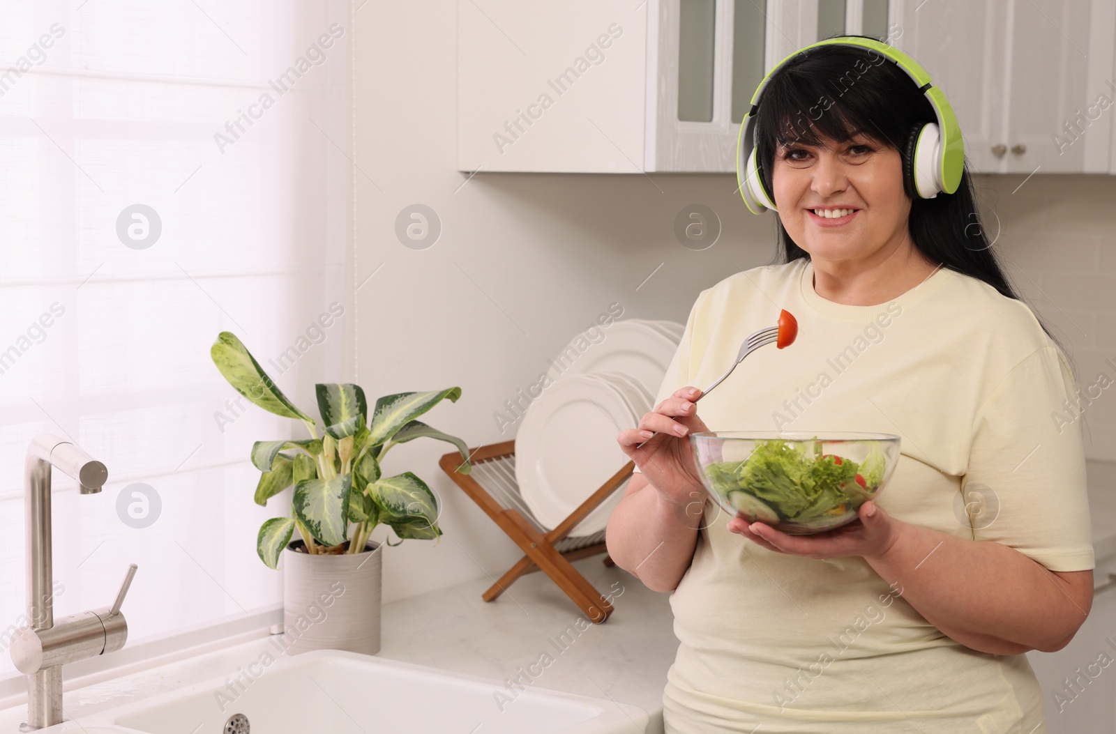 Photo of Happy overweight woman with headphones and bowl of salad in kitchen. Healthy diet