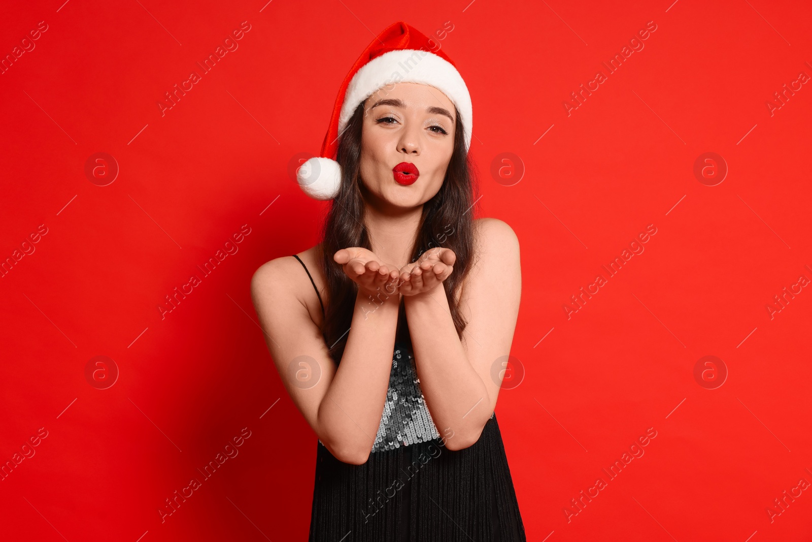 Photo of Christmas celebration. Beautiful young woman in stylish dress and Santa hat blowing kiss on red background