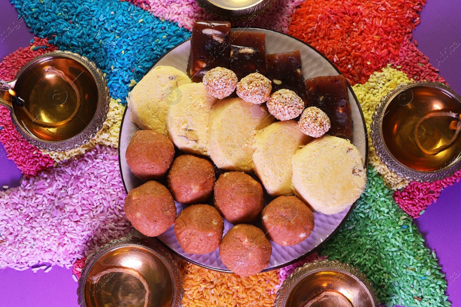 Photo of Diwali celebration. Tasty Indian sweets, diya lamps and colorful rangoli on violet table, flat lay