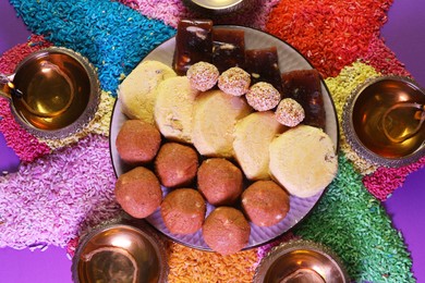 Photo of Diwali celebration. Tasty Indian sweets, diya lamps and colorful rangoli on violet table, flat lay