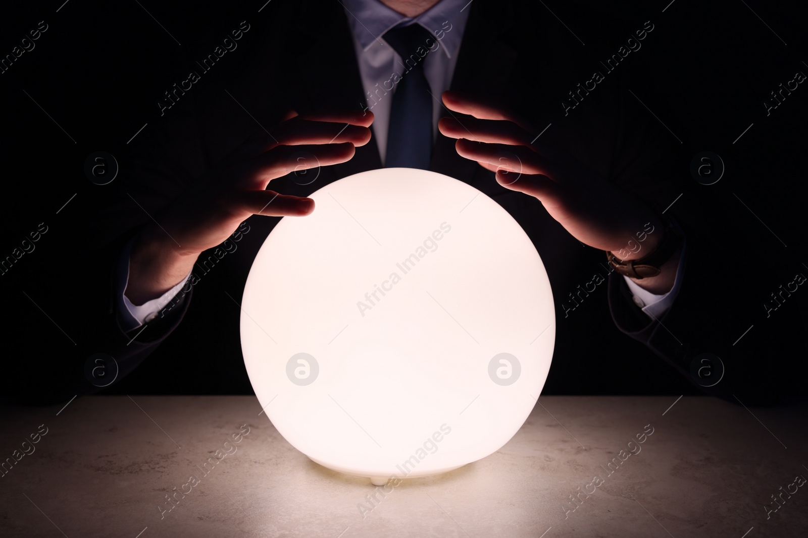 Photo of Businessman using glowing crystal ball to predict future at table in darkness, closeup. Fortune telling