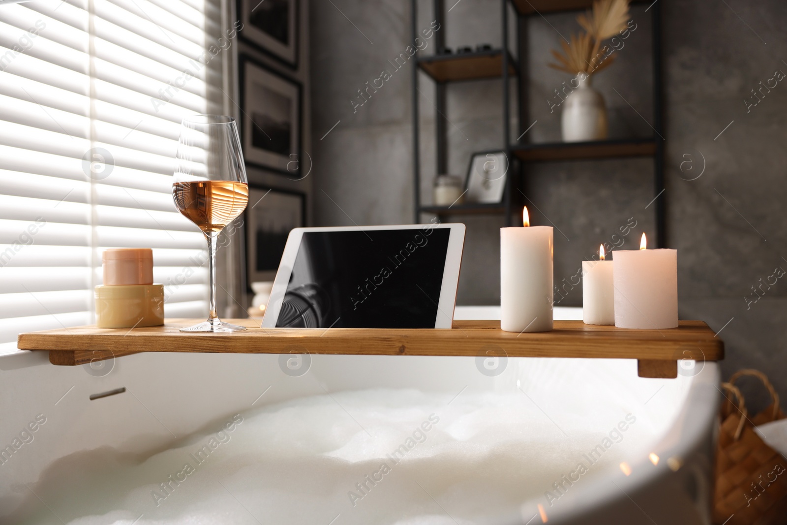 Photo of Wooden tray with tablet, wine, candles and toiletries on bathtub in bathroom