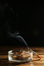Photo of Glass ashtray with stubs and smoldering cigarette on wooden table against black background. Space for text