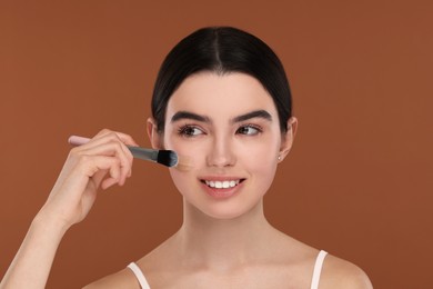 Photo of Teenage girl applying foundation on face with brush against brown background