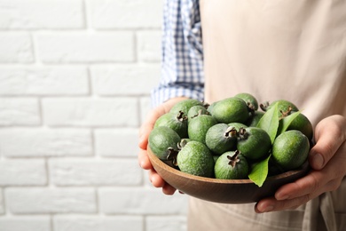 Photo of Woman holding fresh green feijoa fruits against white brick wall, closeup. Space for text
