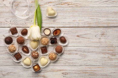 Heart made with delicious chocolate candies and beautiful tulip on white wooden table, flat lay. Space for text