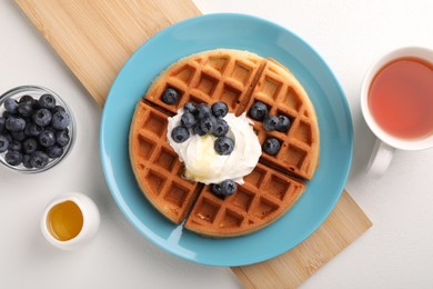 Tasty Belgian waffle with blueberries, honey and whipped cream served on white table, flat lay