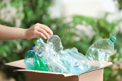 Photo of Woman putting used plastic bottle into cardboard box on blurred background, closeup. Recycling problem