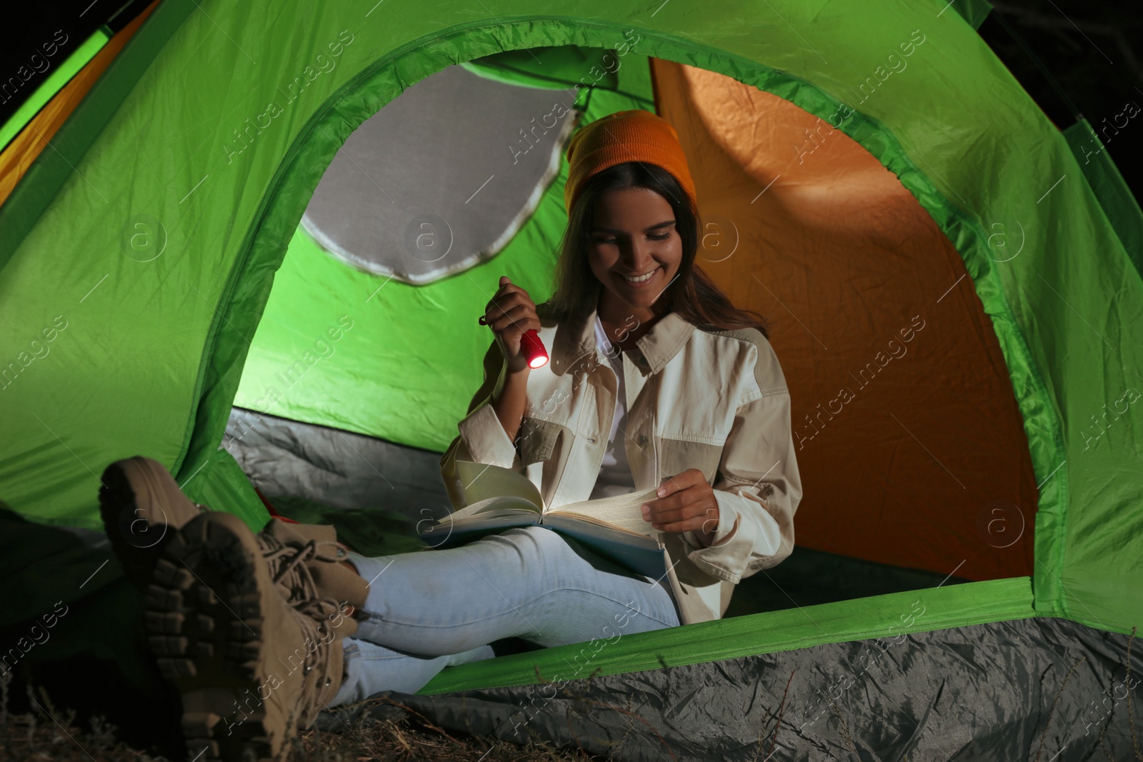 Photo of Young woman with flashlight reading book in tent at night