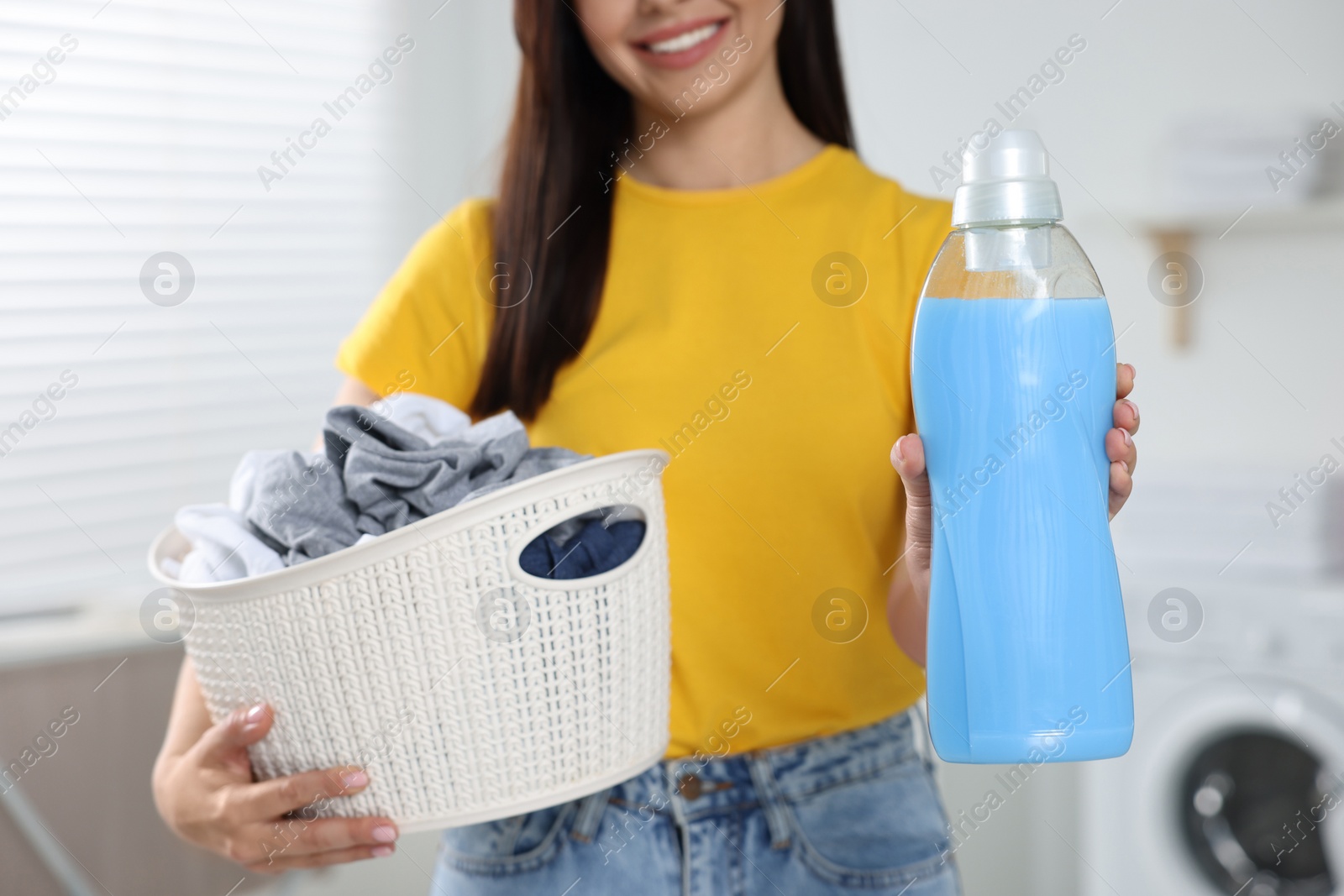 Photo of Woman holding fabric softener and basket with dirty clothes in bathroom, closeup