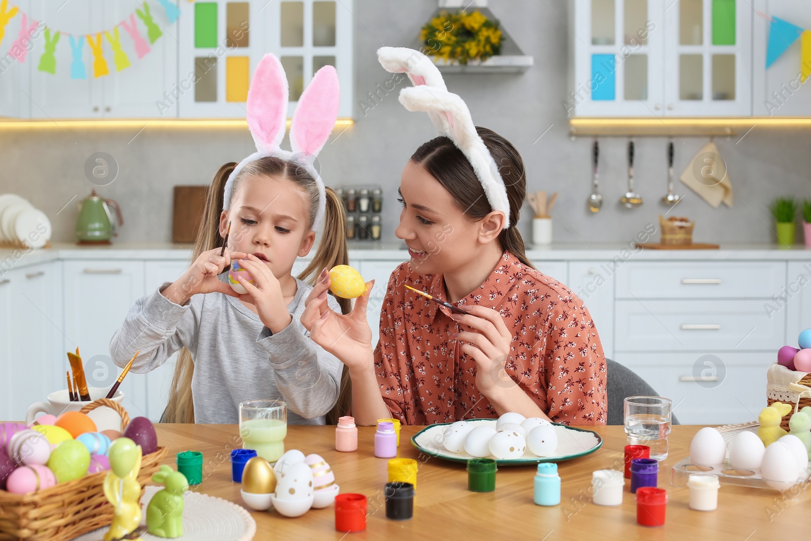 Photo of Mother and her cute daughter painting Easter eggs at table in kitchen