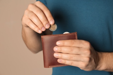 Young man putting coin into wallet on beige background, closeup