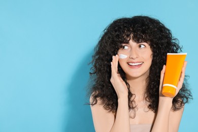 Photo of Beautiful young woman with tube of sunscreen applying sun protection cream onto her face on light blue background, space for text