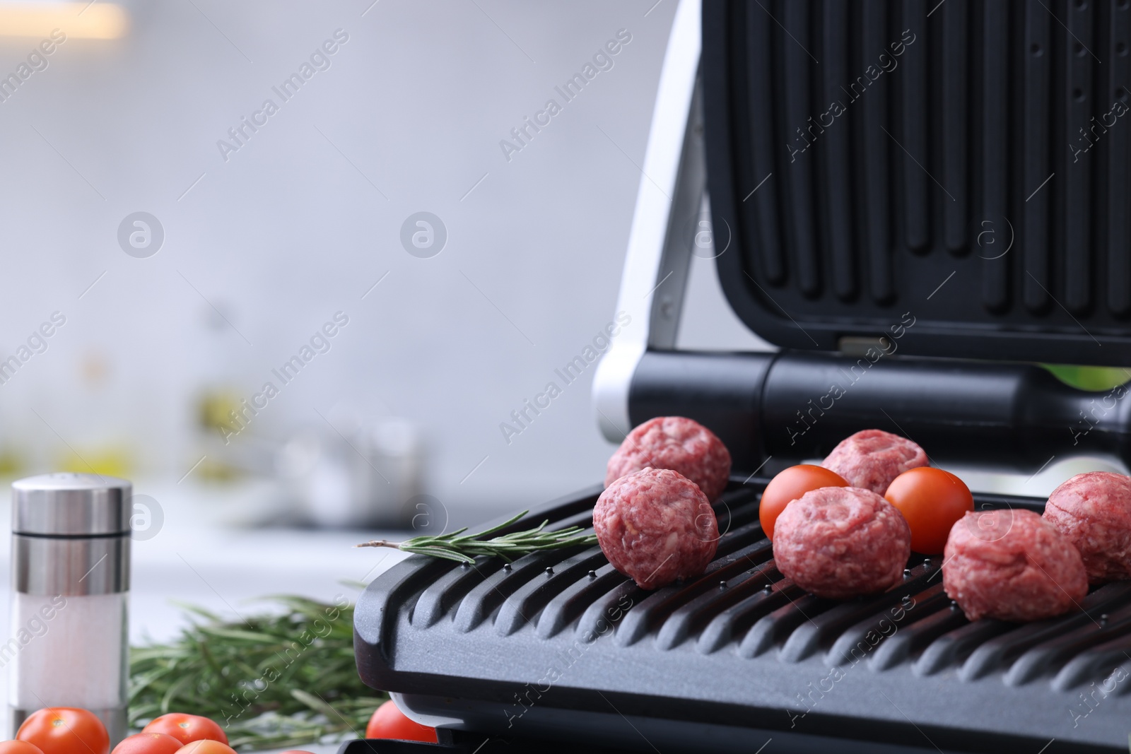 Photo of Electric grill with meatballs, tomatoes and rosemary on table against blurred background, closeup