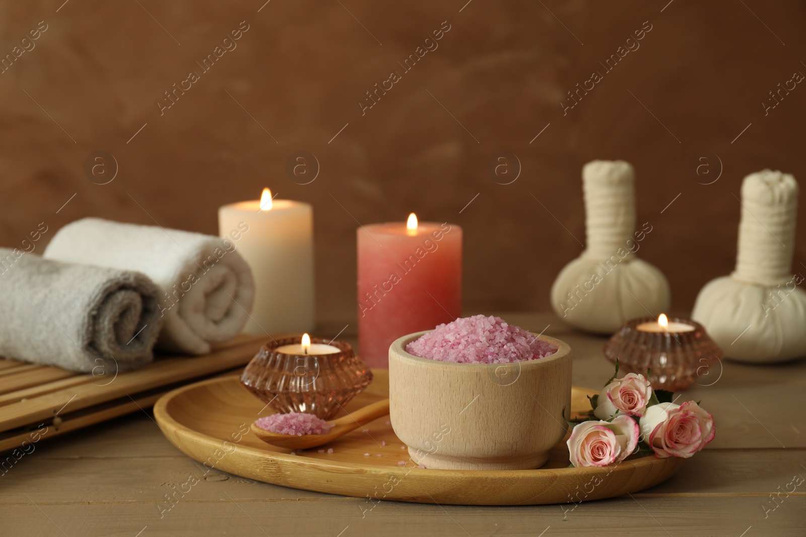 Photo of Bowl of pink sea salt, roses, burning candles, herbal massage bags and towels on wooden table