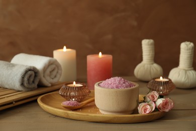 Bowl of pink sea salt, roses, burning candles, herbal massage bags and towels on wooden table