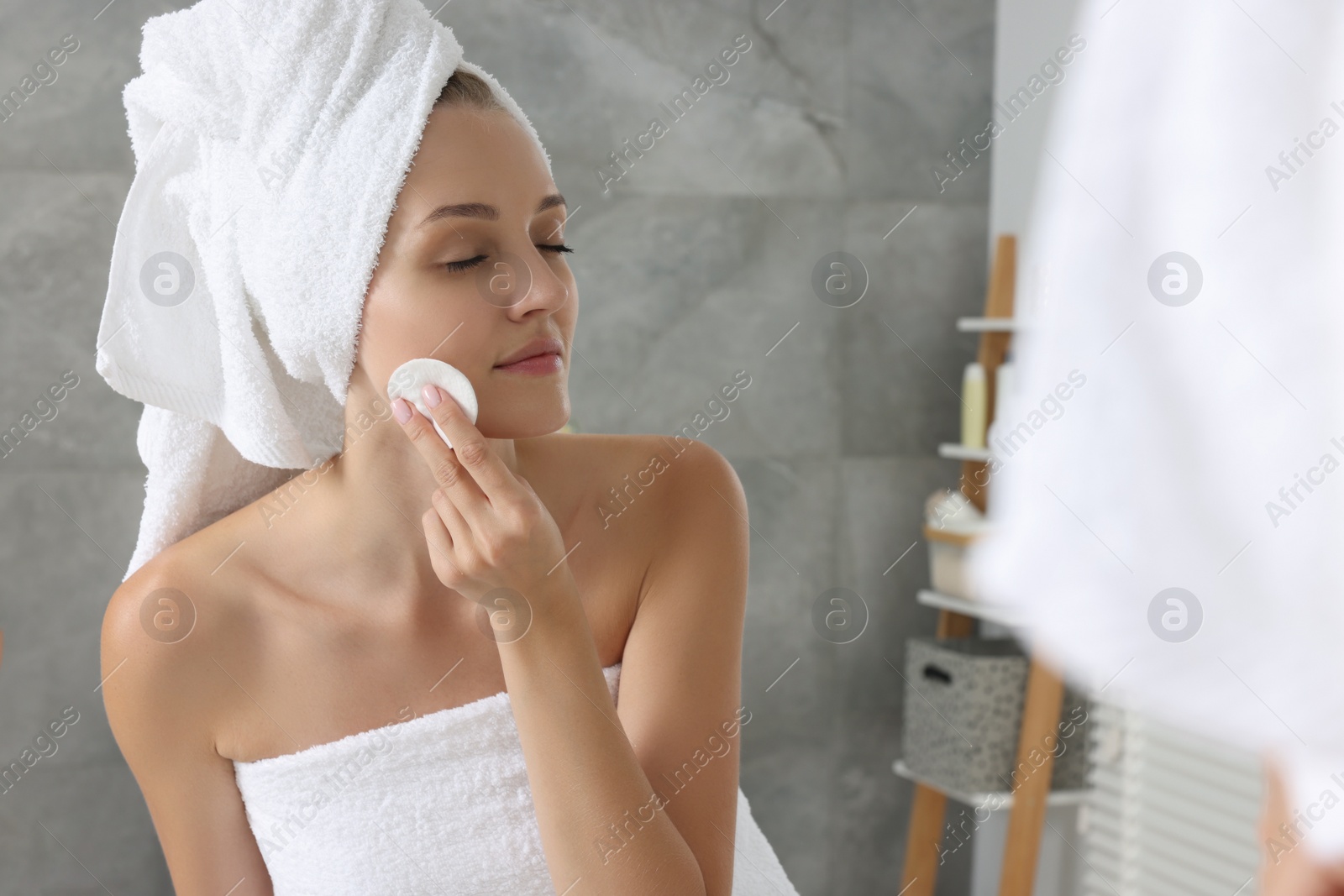 Photo of Young woman cleaning her face with cotton pad near mirror in bathroom