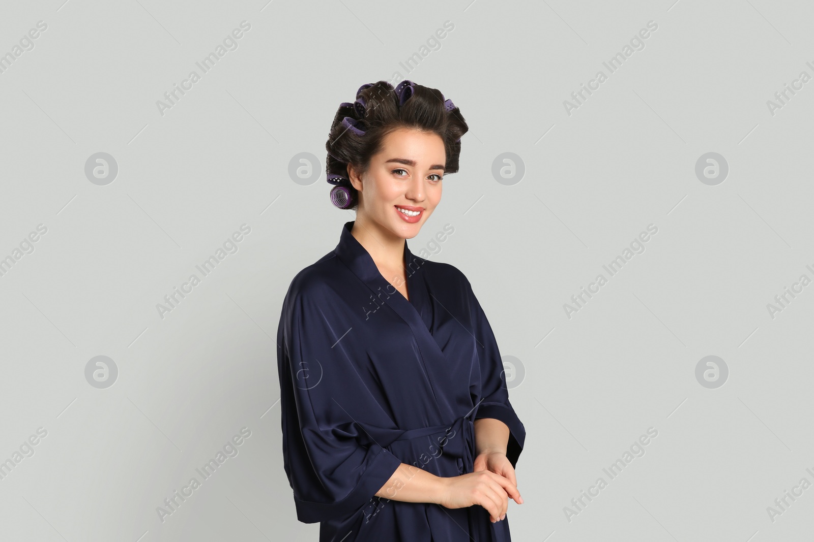 Photo of Happy young woman in silk bathrobe with hair curlers on grey background