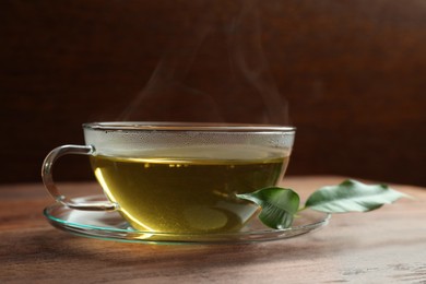 Photo of Fresh green tea in glass cup with saucer and leaves on wooden table