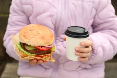 Little girl holding fresh delicious burger and cup of coffee outdoors, closeup. Street food