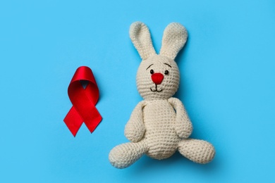 Photo of Cute knitted toy bunny and red ribbon on blue background, flat lay. AIDS disease awareness