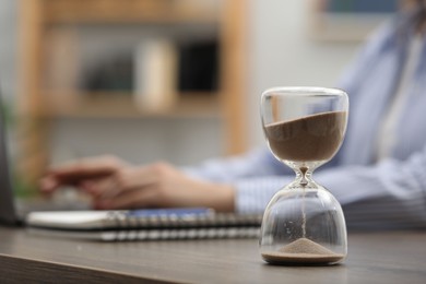 Photo of Hourglass with flowing sand on desk. Woman using laptop indoors, selective focus