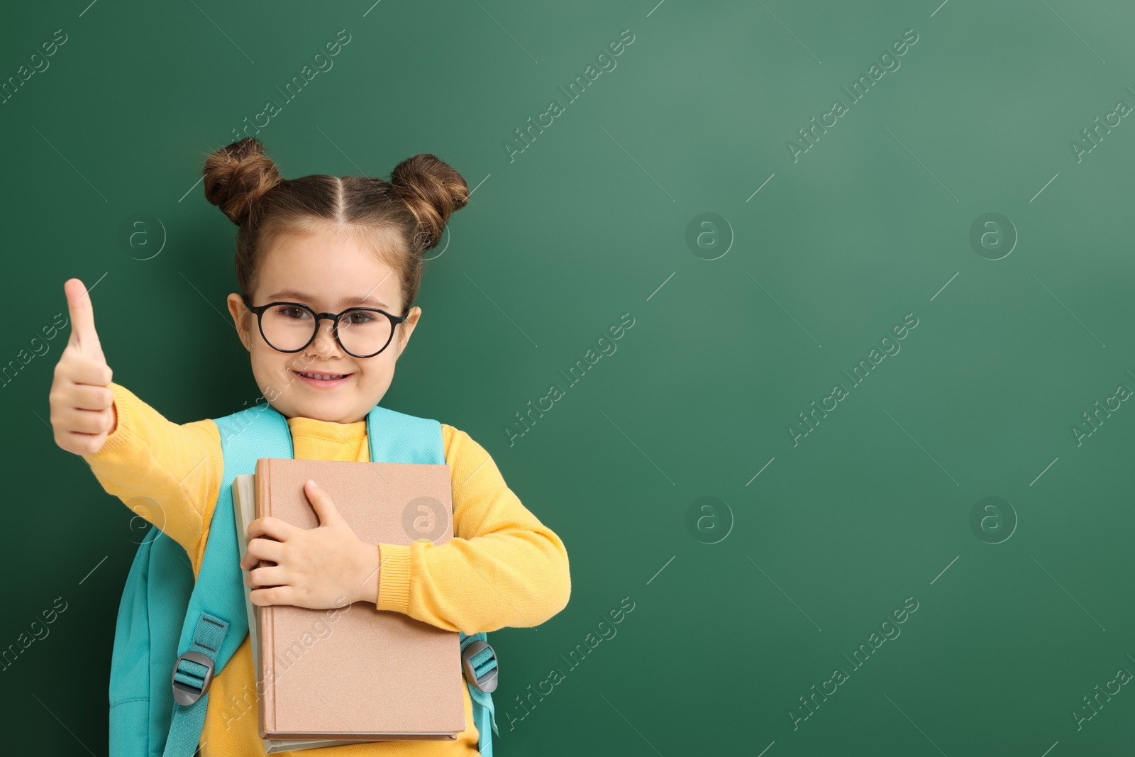 Photo of Happy little school child with notebooks showing thumbs up near chalkboard. Space for text