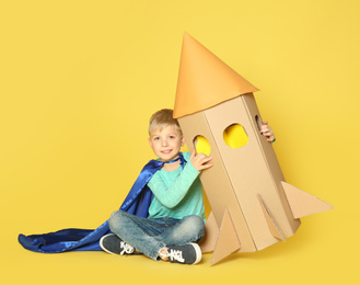 Photo of Little child in cape playing with rocket made of cardboard box on yellow background