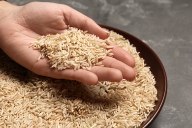 Woman holding grains near plate with brown rice on table, closeup