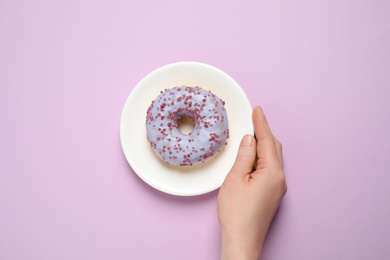 Photo of Woman holding plate with delicious glazed donut on lilac background, top view