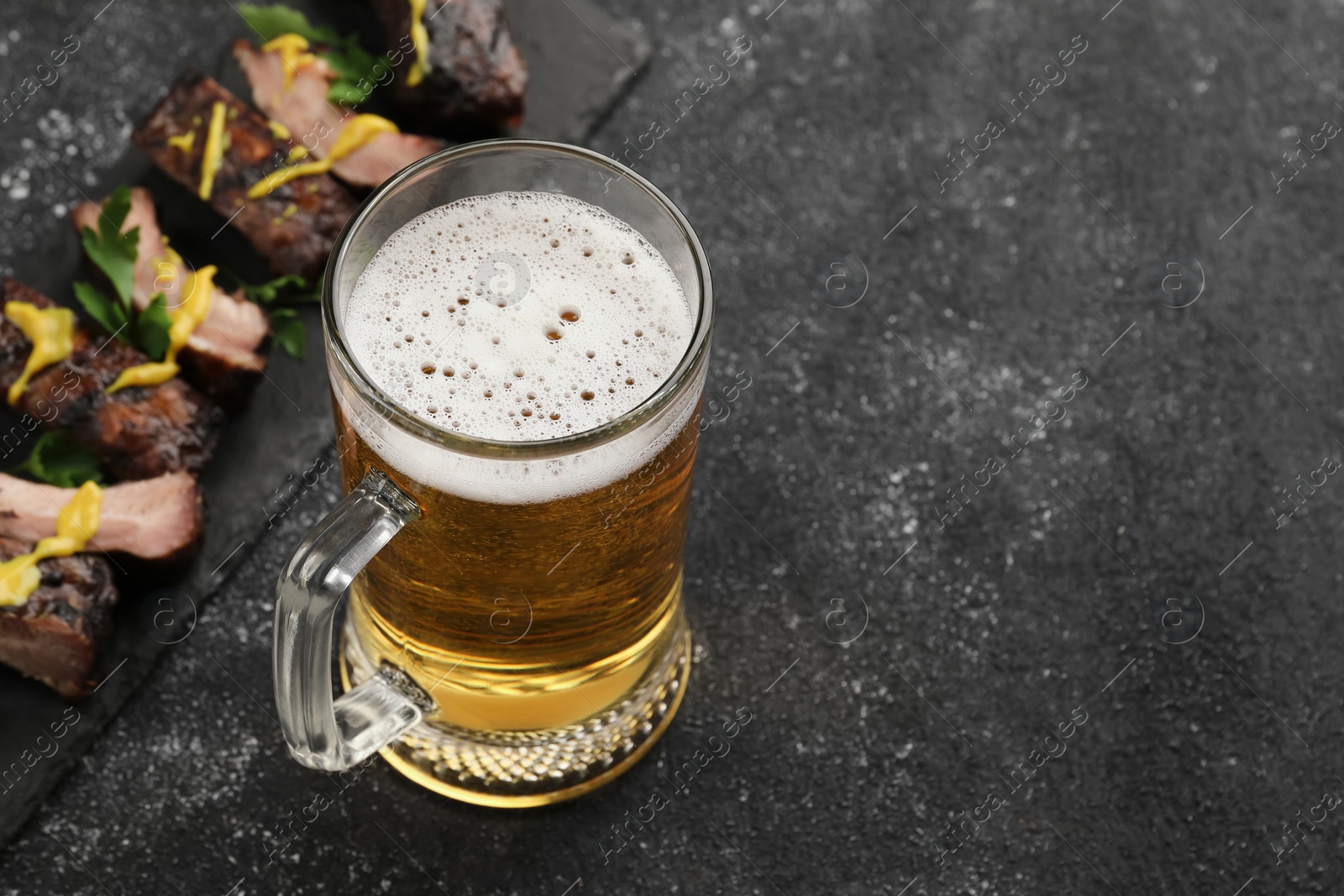 Photo of Mug with beer and delicious grilled ribs on dark grey textured table. Space for text