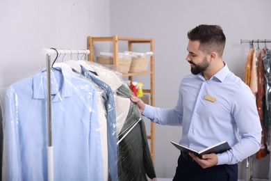 Photo of Dry-cleaning service. Happy worker with notebook taking coat from rack indoors