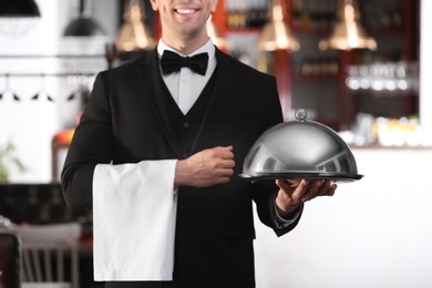 Photo of Waiter in elegant uniform holding metal tray and cloche at workplace