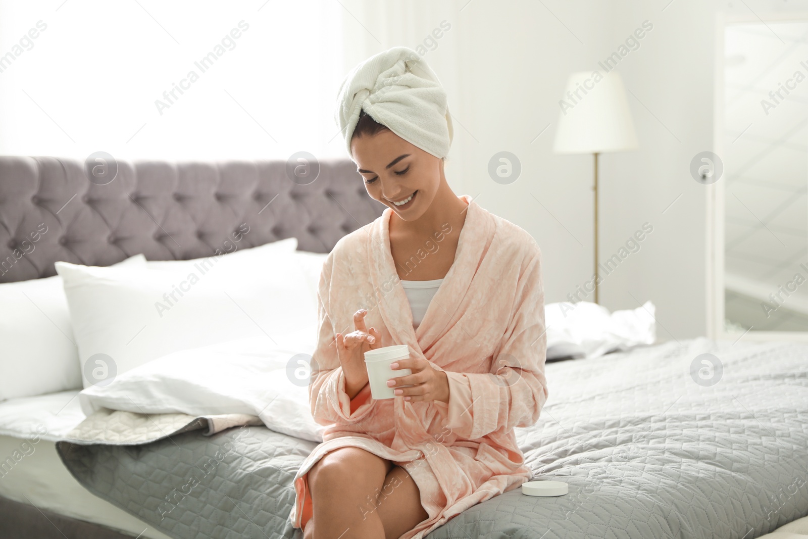 Photo of Pretty young woman with towel on head using skin cream in bedroom