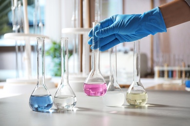 Photo of Scientist taking flask with colorful liquid from table indoors, closeup. Laboratory analysis