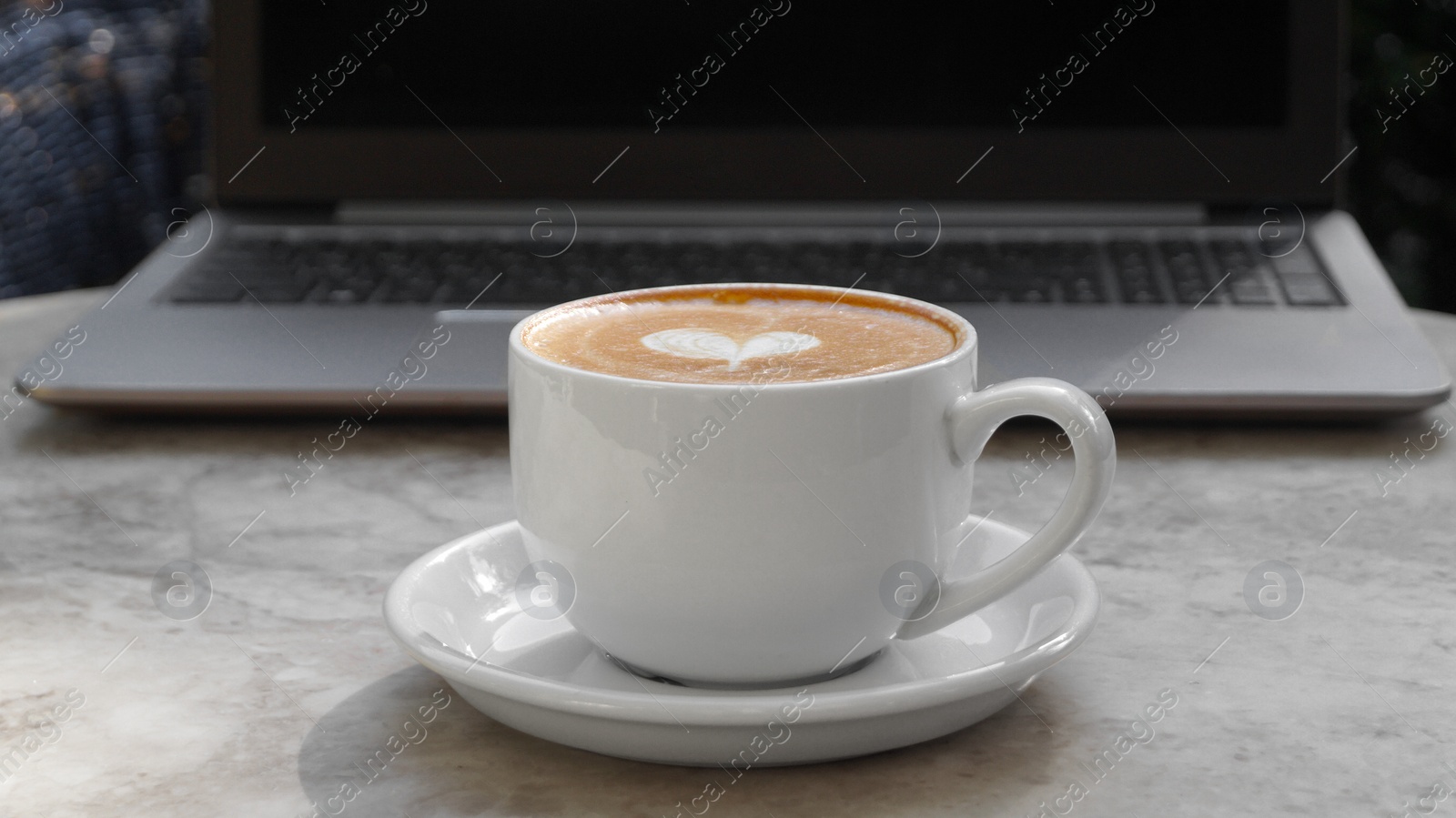 Photo of Cup of delicious coffee and laptop on beige marble table outdoors