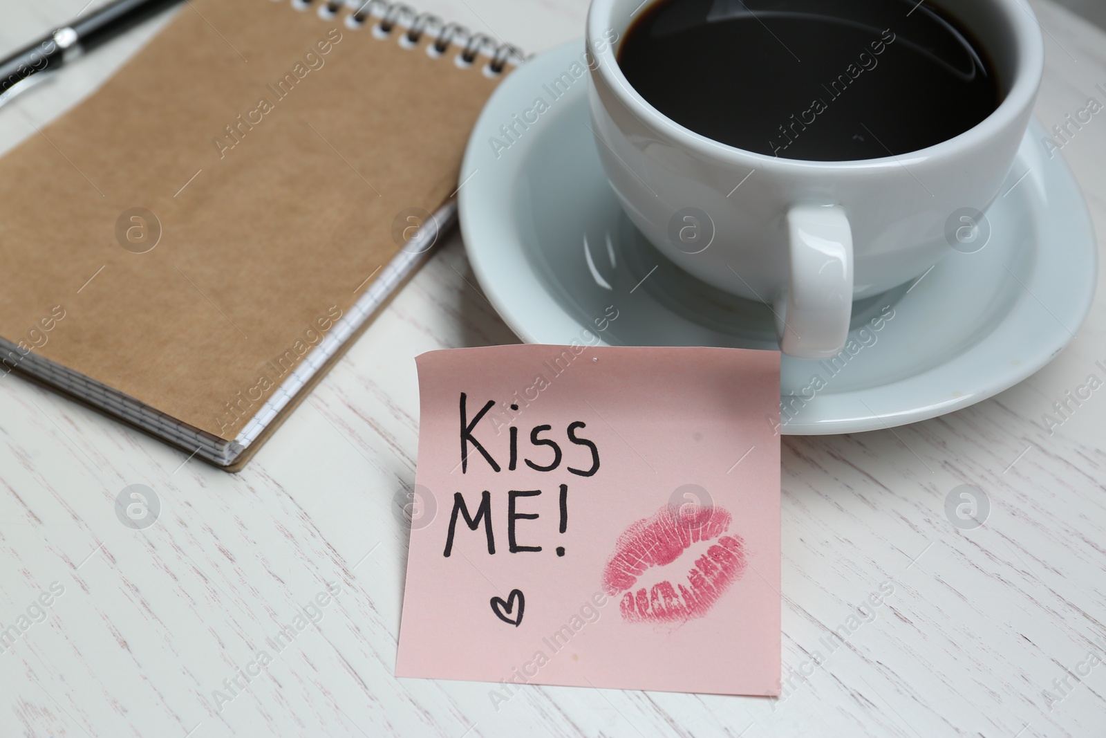 Photo of Sticky note with phrase Kiss Me, lipstick mark, cup of drink and notebook on white wooden table