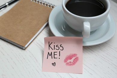 Sticky note with phrase Kiss Me, lipstick mark, cup of drink and notebook on white wooden table