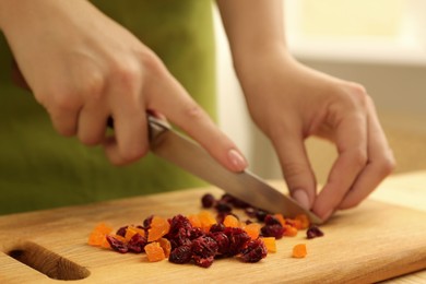 Photo of Making granola. Woman cutting dried apricots and cherries at wooden board in kitchen, closeup