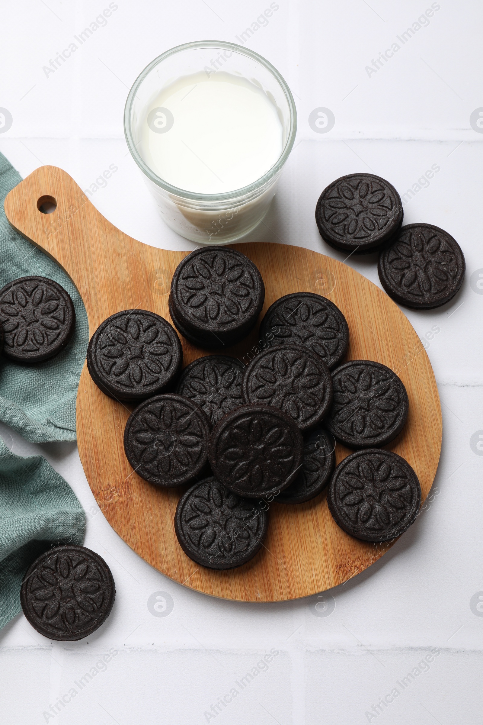 Photo of Board with tasty sandwich cookies and milk on white tiled table, flat lay