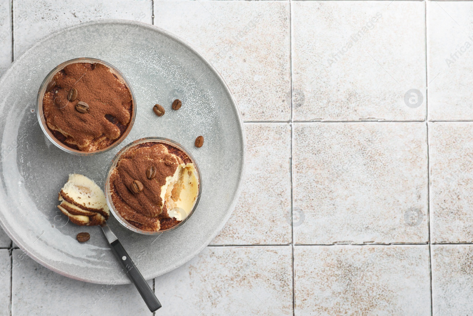 Photo of Delicious tiramisu in glasses, spoon and coffee beans on light tiled table, top view. Space for text