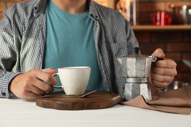 Brewing coffee. Man with moka pot and cup at white wooden table indoors, closeup