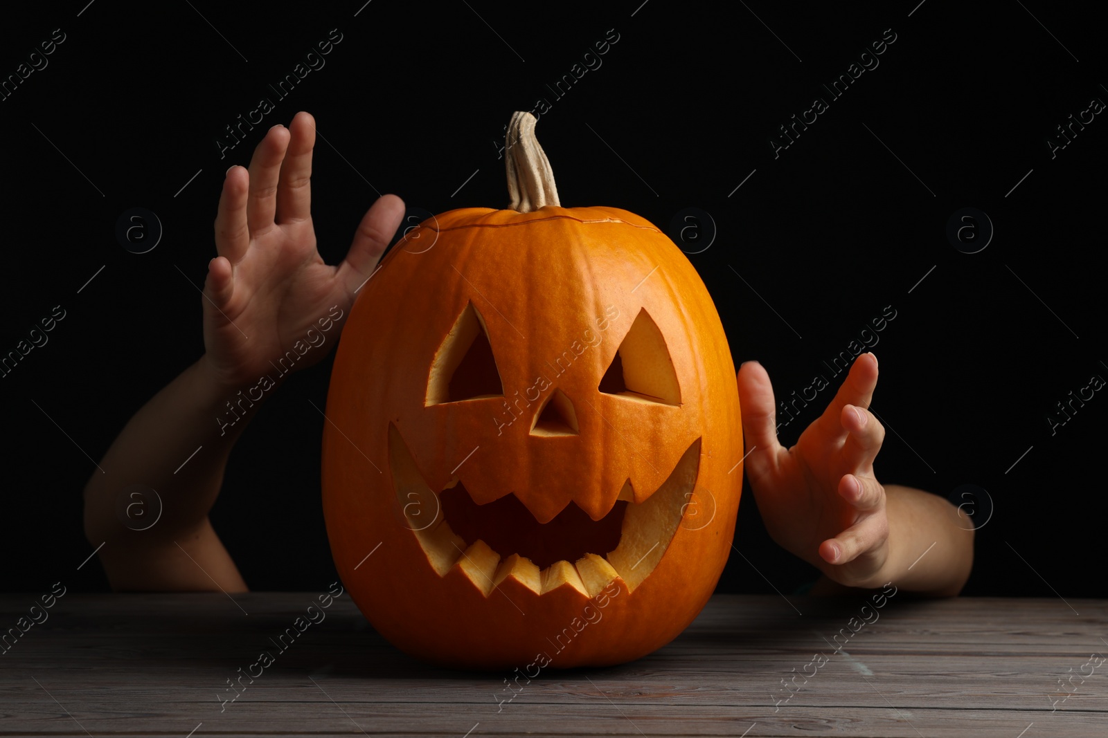 Photo of Woman with scary jack o'lantern made of pumpkin on wooden table against black background, closeup. Halloween traditional decor