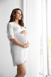 Young pregnant woman in lace nightgown near window indoors. Space for text