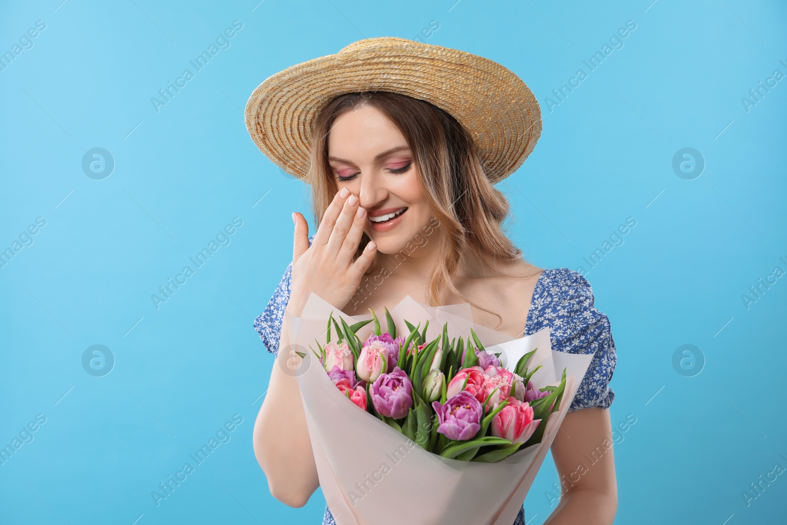 Photo of Happy young woman in straw hat holding bouquet of beautiful tulips on light blue background