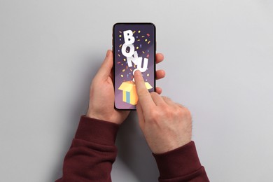 Image of Bonus gaining. Man using smartphone on light grey background, top view. Illustration of open gift box, word and confetti on device screen