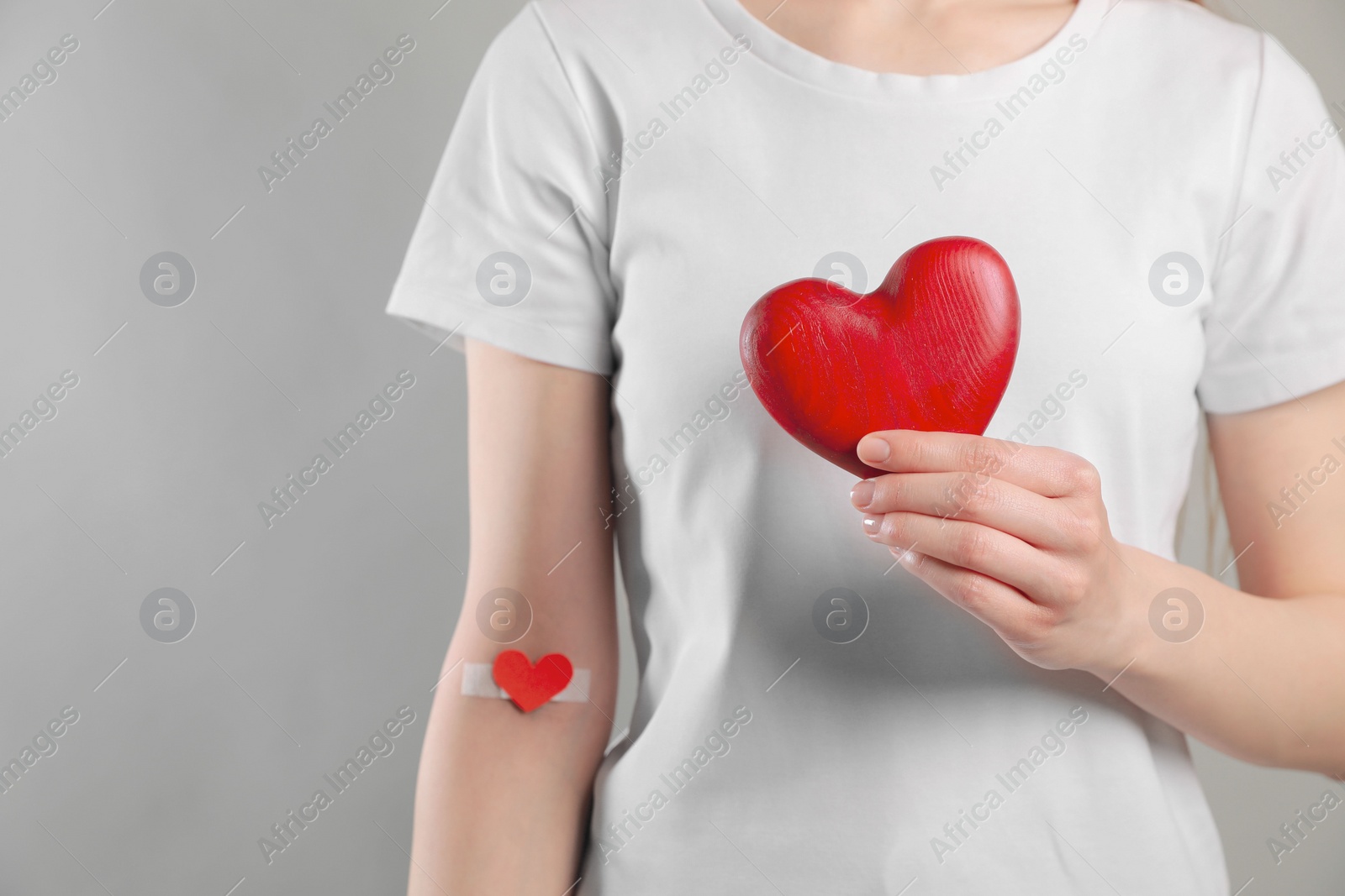 Photo of Blood donation concept. Woman with adhesive plaster on arm holding red heart against grey background, closeup. Space for text