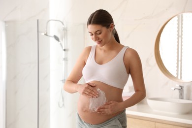 Photo of Young pregnant woman applying cosmetic product on belly in bathroom